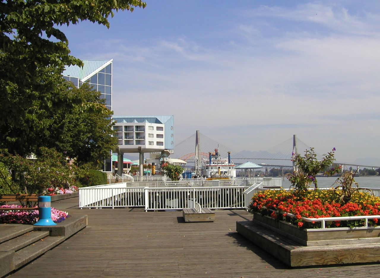 Sunlit flowers and the boardwalk along New Westminster Quay, with the Fraser River beyond